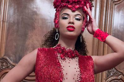 Femme portant une tenue rouge ornée de perles et un accessoire de tête assorti, posant devant une porte en bois sculpté.