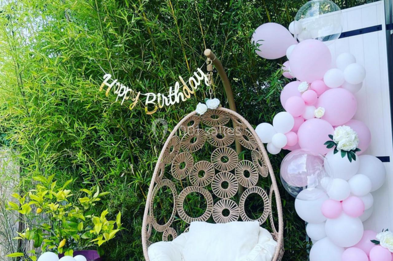 Fauteuil suspendu en rotin avec coussin blanc, entouré de ballons roses et blancs, devant un fond de bambous.