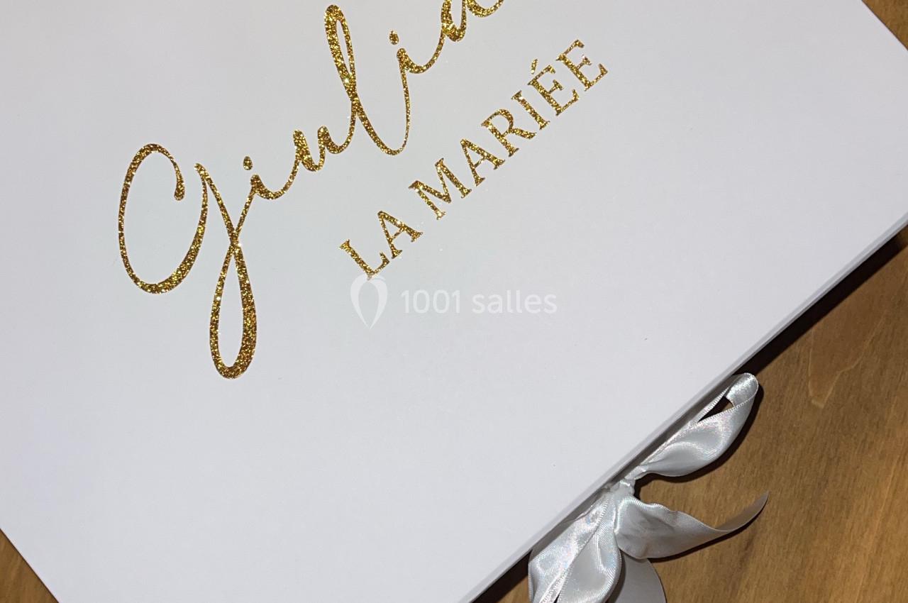 Boîte blanche avec l'inscription dorée ’Giulia La Mariée’ posée sur une table en bois, fermée par un ruban blanc.