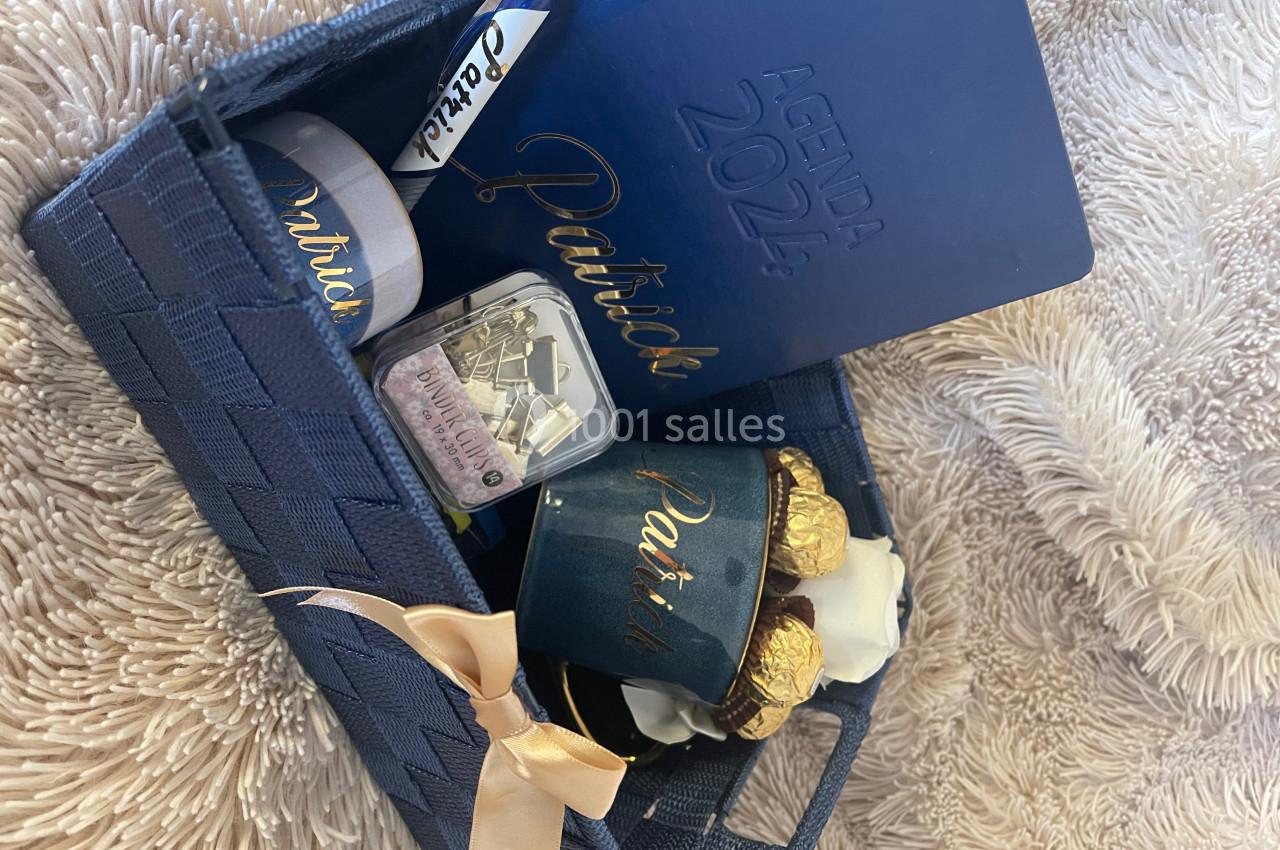 Panier cadeau bleu contenant un agenda, une tasse, des chocolats et une petite boîte, posé sur un tissu beige.