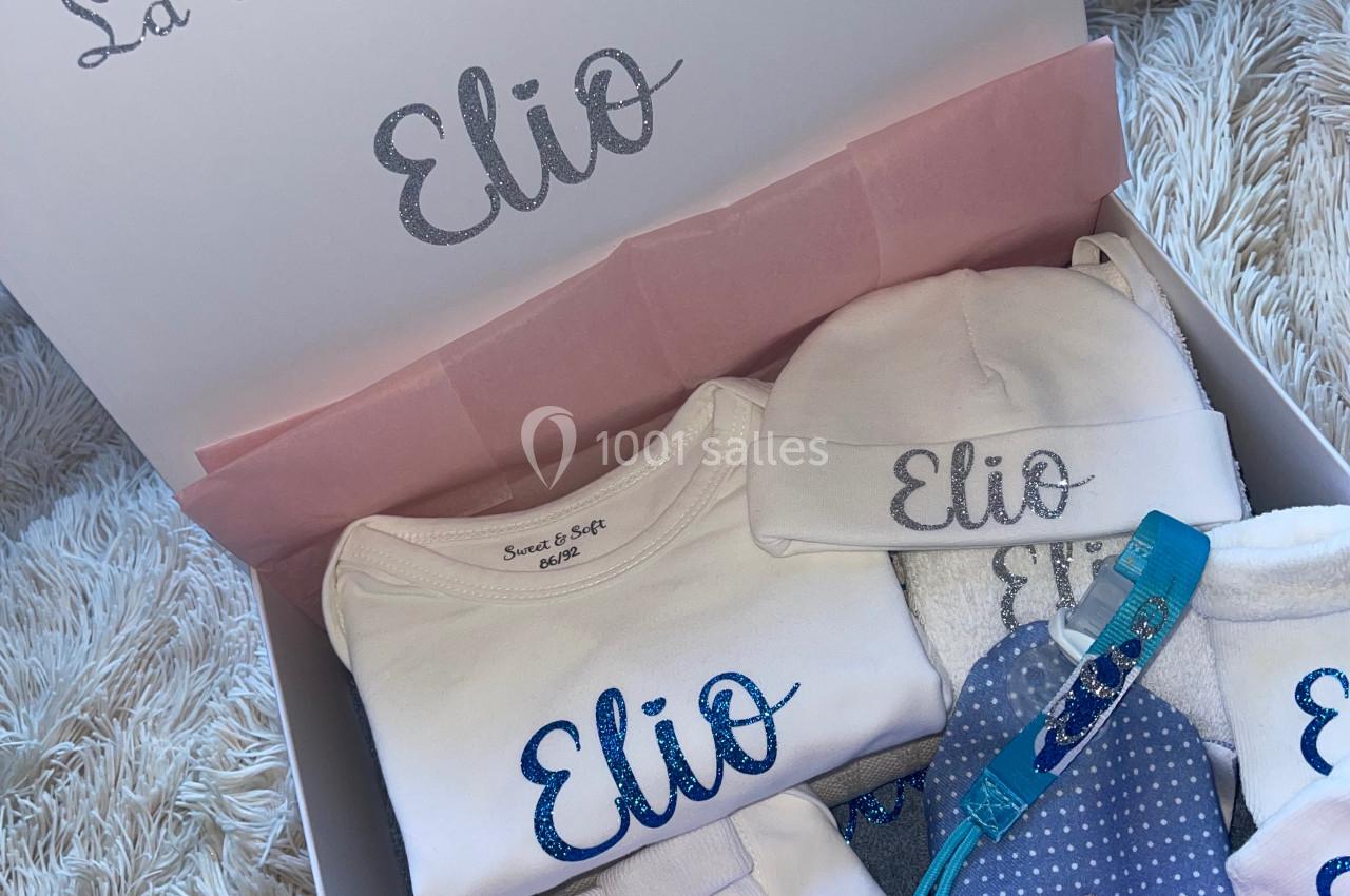 Boîte contenant des vêtements et accessoires pour bébé personnalisés avec le prénom ’Elio’, présentés sur un fond doux.