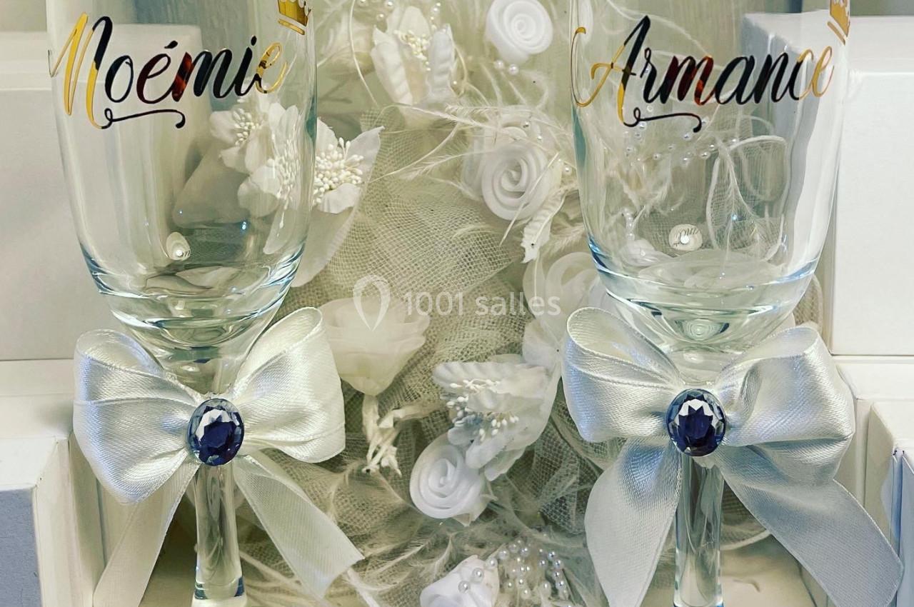 Deux flûtes à champagne personnalisées avec des prénoms, ornées de nœuds blancs et posées devant une décoration florale.