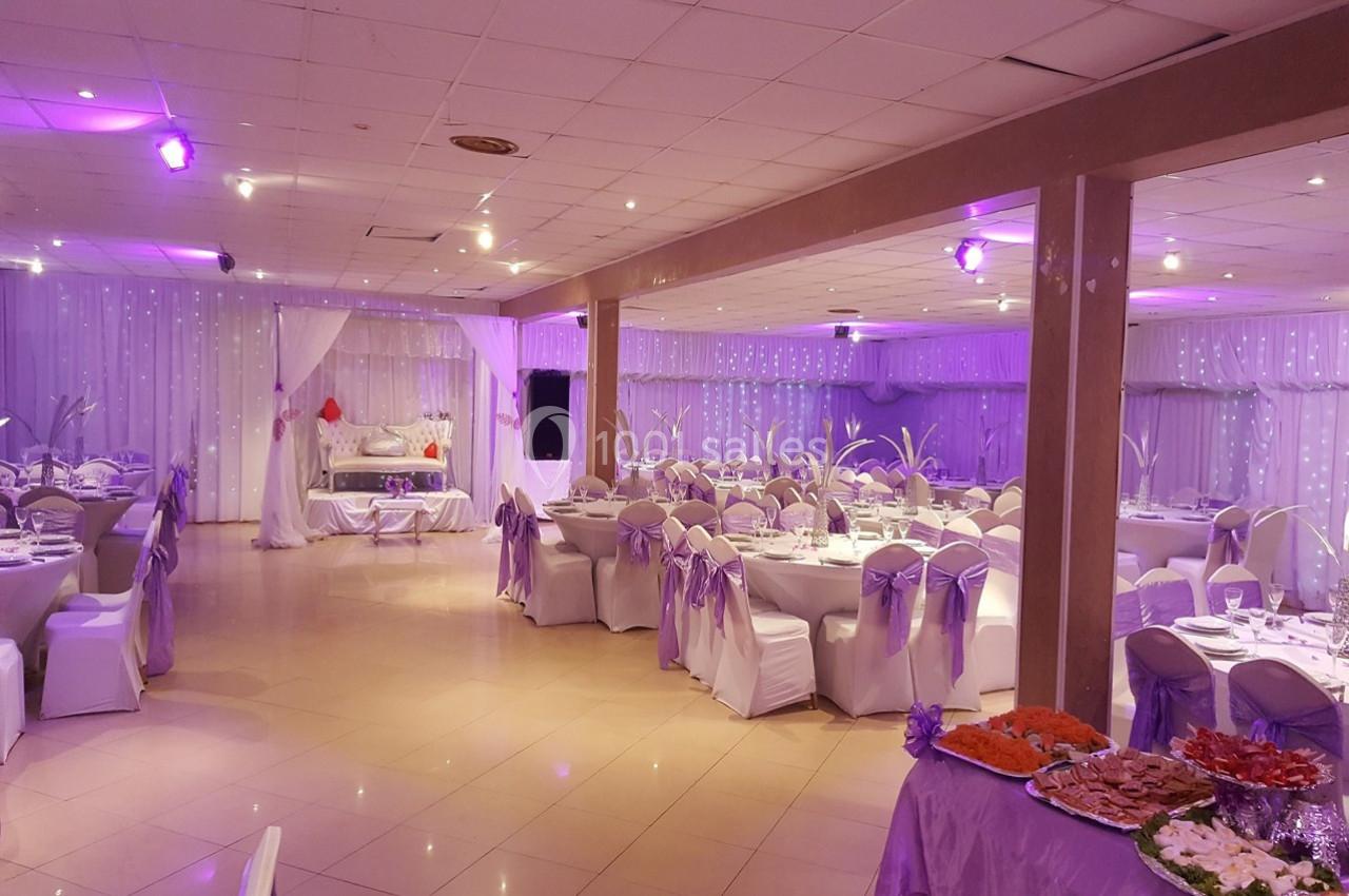 Salle de réception décorée avec des nappes et chaises blanches ornées de rubans violets, éclairée par une lumière tamisée.