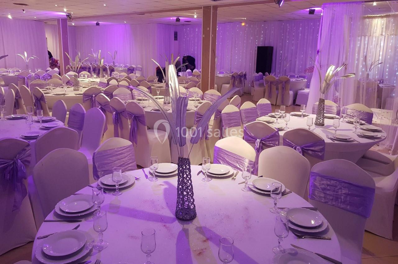 Salle de réception décorée avec des tables rondes, nappes blanches, chaises ornées de rubans violets et éclairage tamisé.