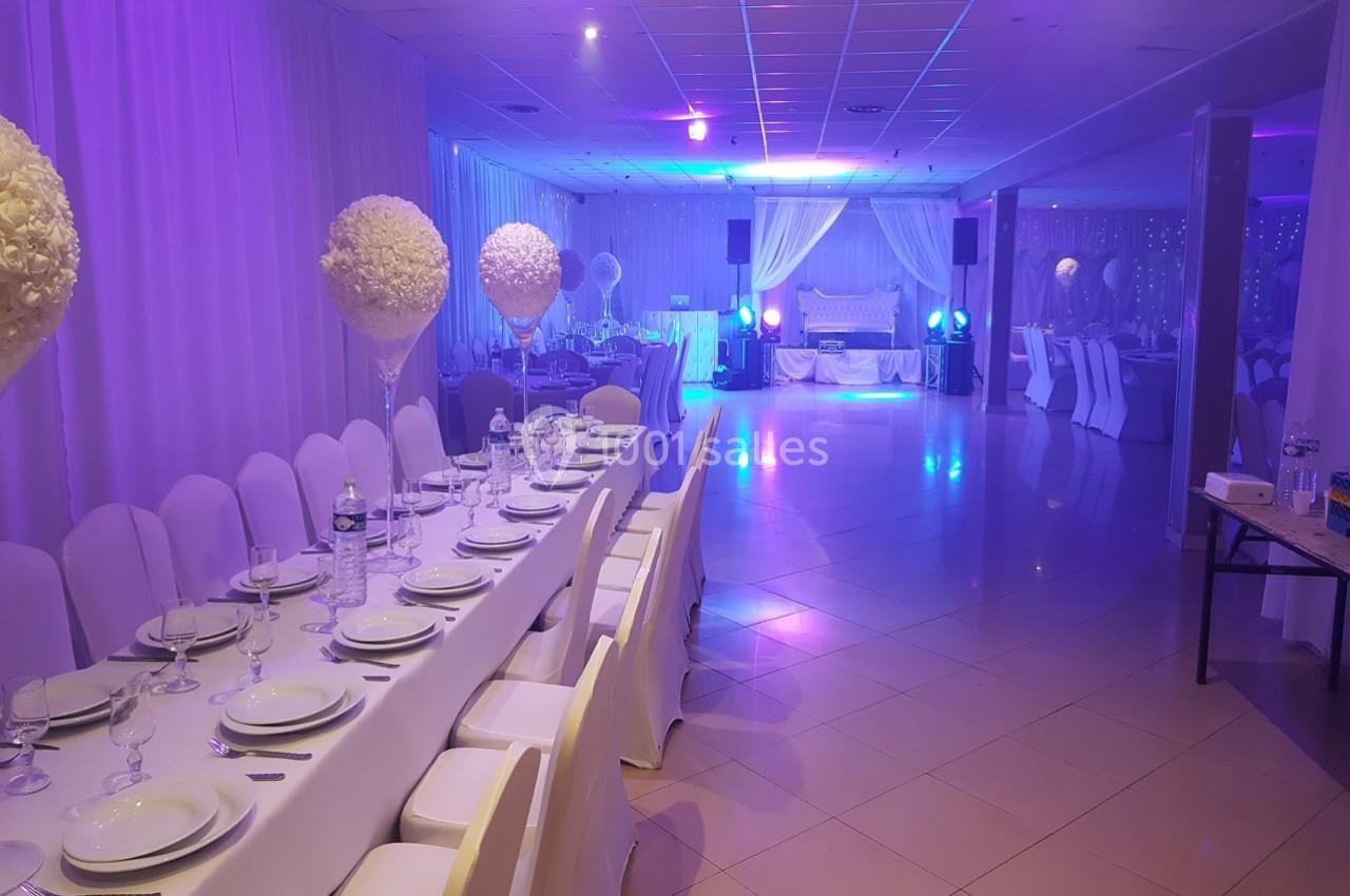 Salle de réception décorée avec des tables dressées, des chaises blanches et un éclairage tamisé bleu et violet.