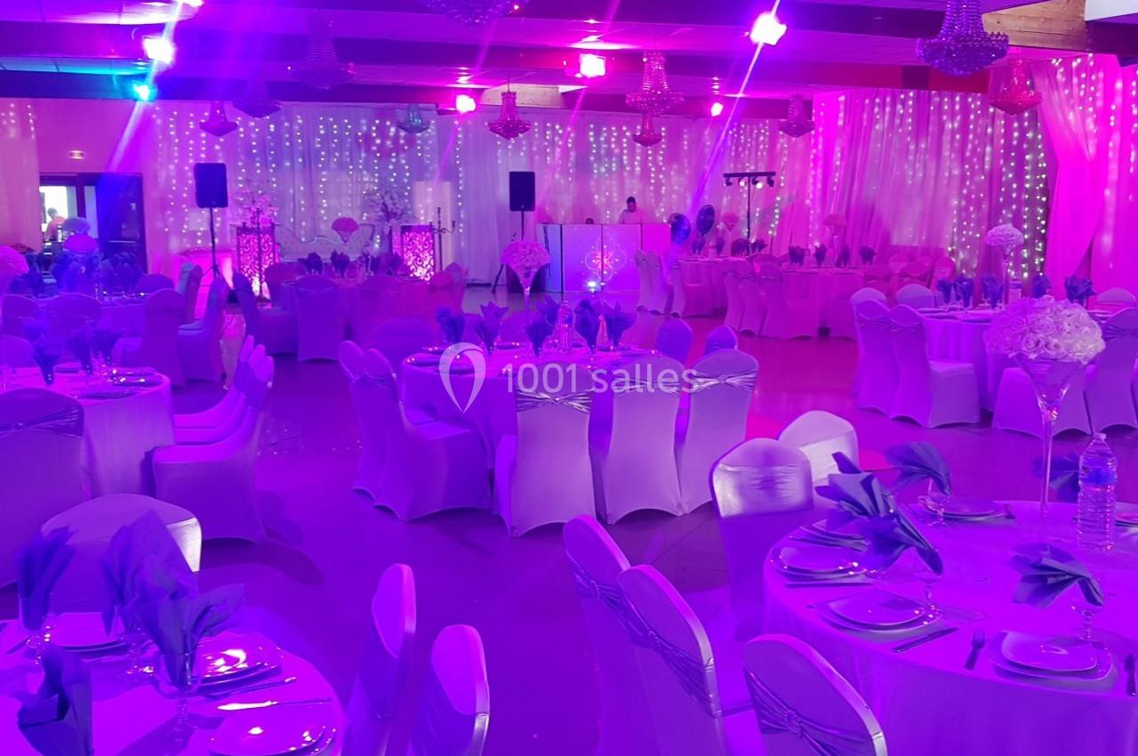 Salle de réception décorée avec des tables rondes, nappes blanches, serviettes violettes et éclairage lumineux coloré.