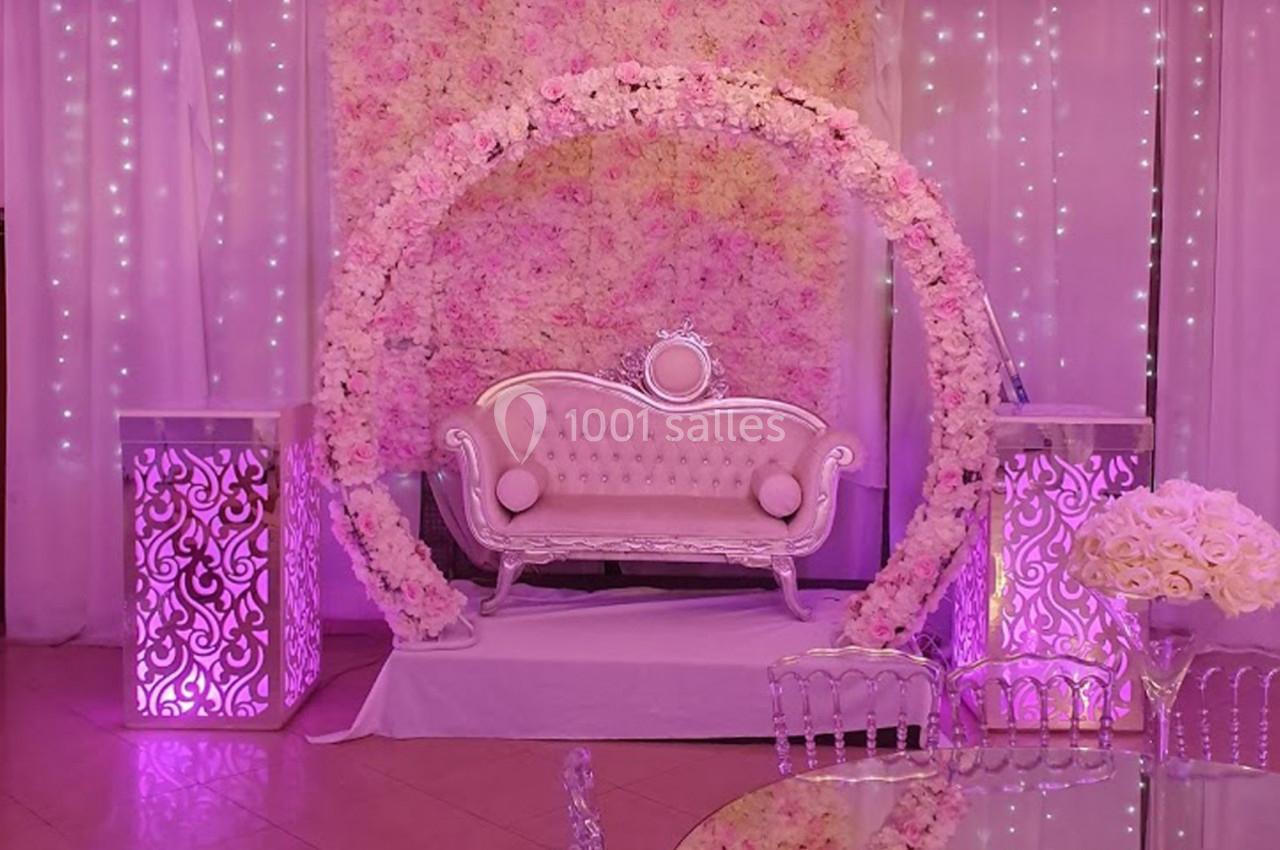 Décor de mariage avec canapé capitonné, arche florale rose, colonnes lumineuses et éclairage violet.