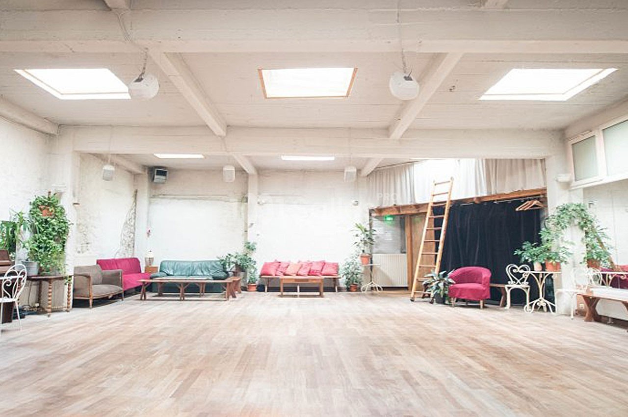 Salle lumineuse avec parquet, canapés colorés, plantes, échelle en bois et rideaux noirs en arrière-plan.