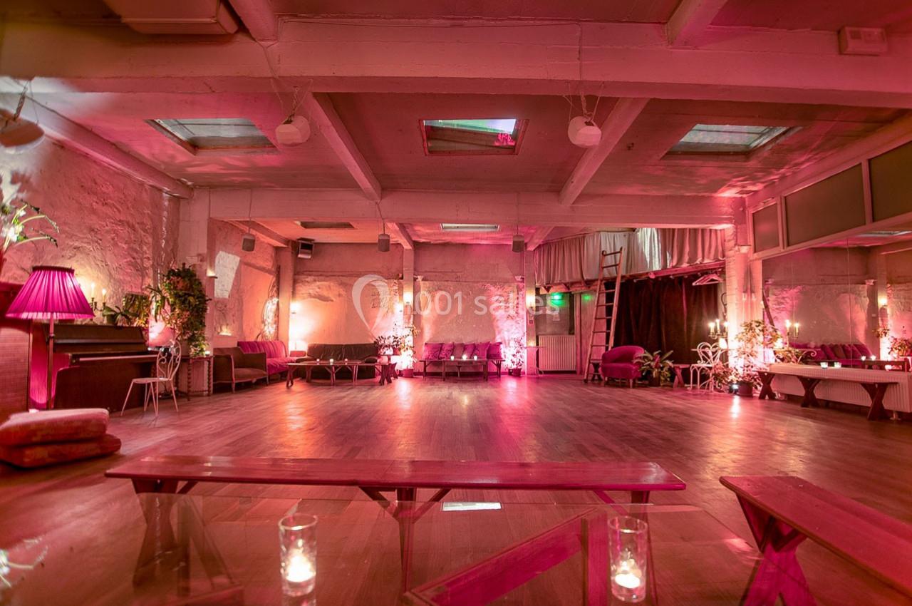 Salle spacieuse avec parquet, éclairage rose tamisé, canapés, plantes et piano dans un décor industriel.