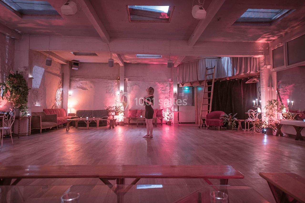 Salle spacieuse avec parquet, éclairage tamisé rose, canapés, plantes et une personne debout au centre.