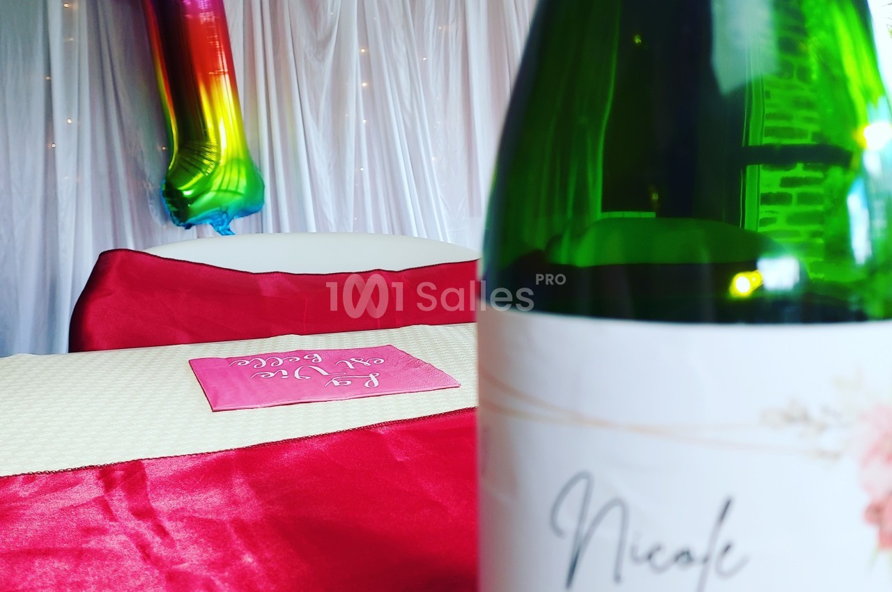 Bouteille de vin au premier plan sur une table rouge, ballon arc-en-ciel en forme de chiffre 1 en arrière-plan.