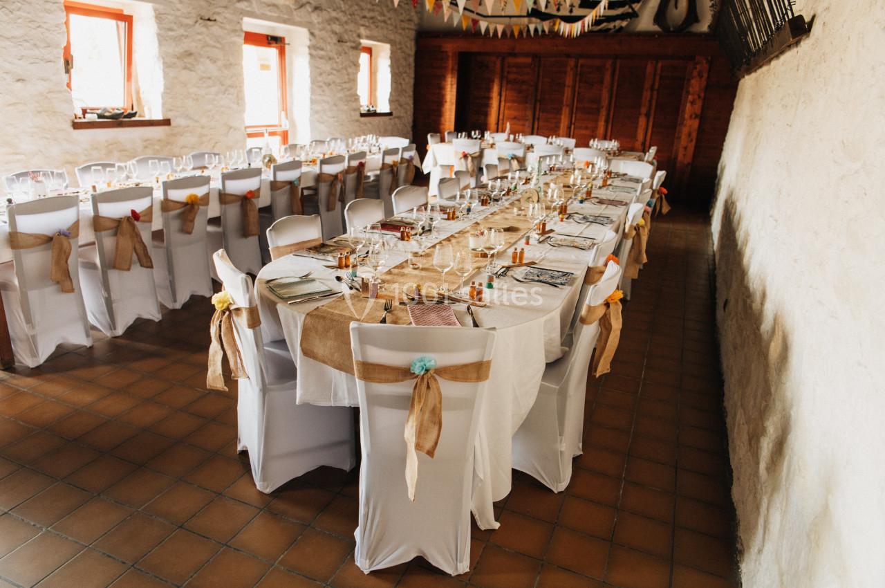 Salle décorée pour un événement avec de longues tables dressées, nappes blanches et chaises ornées de rubans.