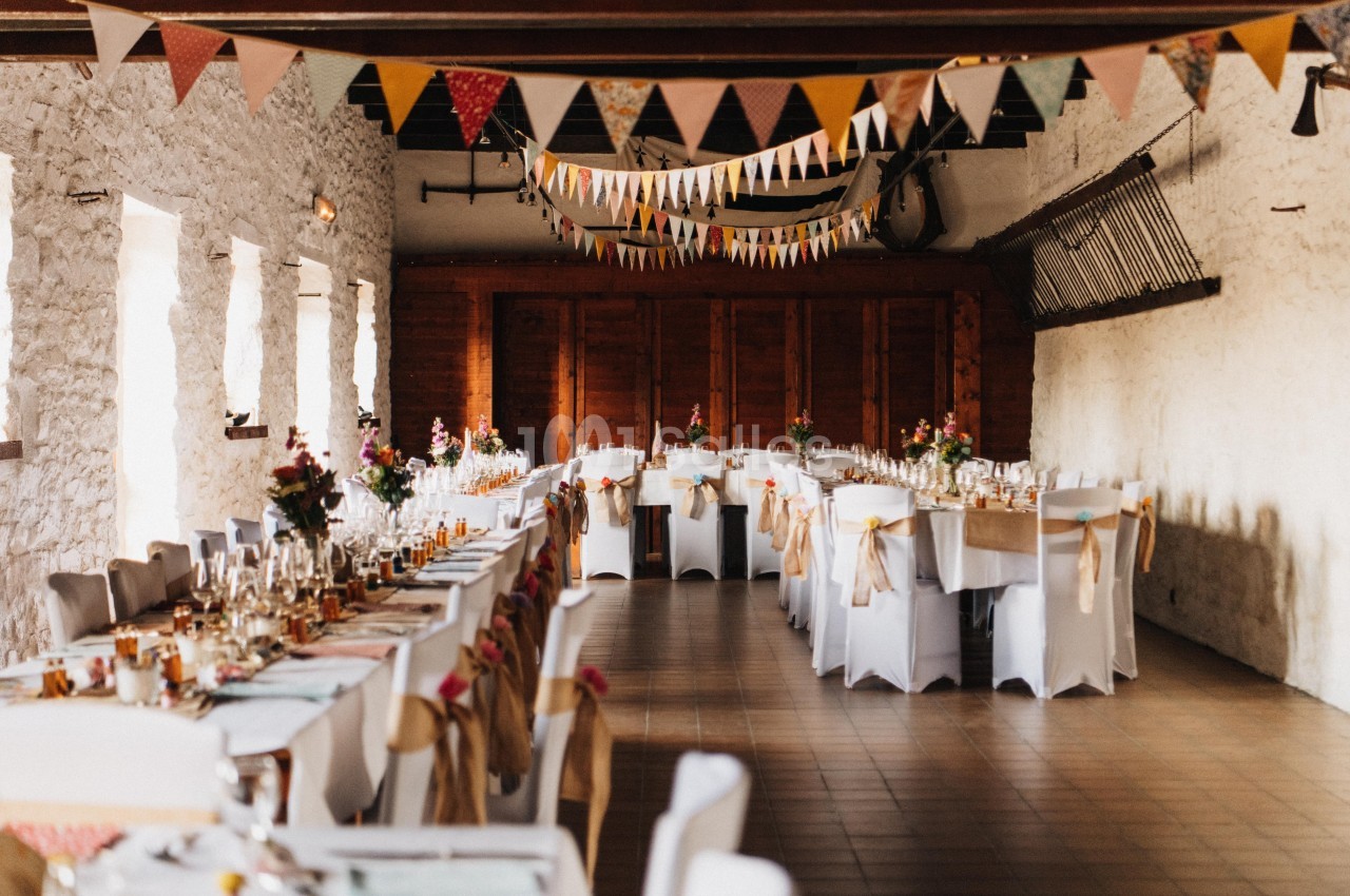 Grande salle décorée pour un événement, avec longues tables dressées, chaises blanches et guirlandes suspendues.
