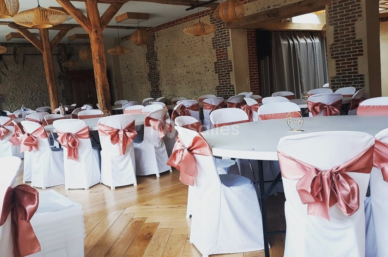 Salle de réception avec des tables rondes et des chaises blanches décorées de nœuds en tissu rose saumon.
