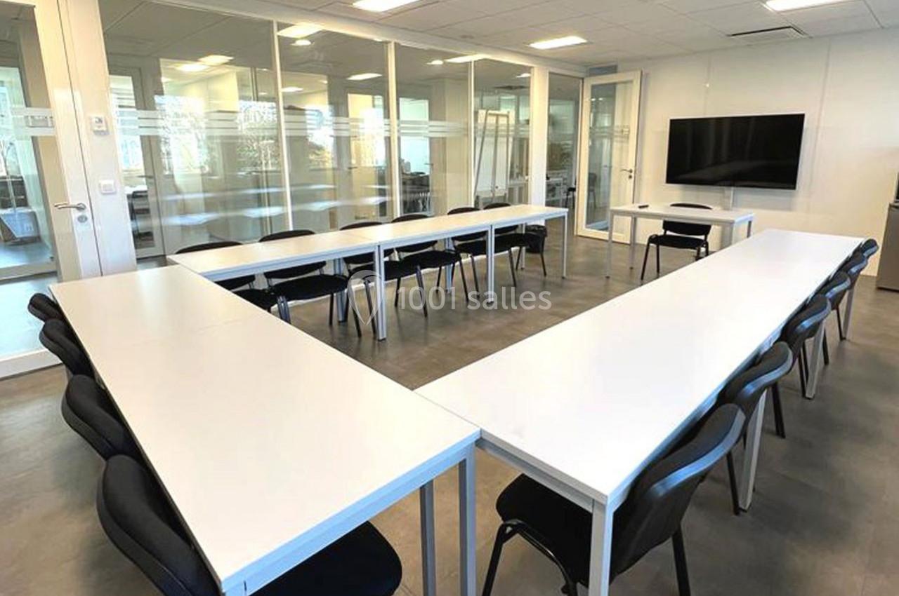 Salle de réunion lumineuse avec des tables blanches disposées en U et des chaises noires, écran mural au fond.
