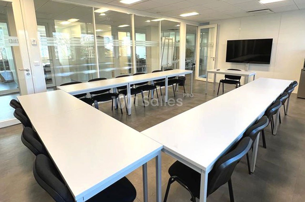 Salle de réunion lumineuse avec des tables blanches disposées en U et des chaises noires, écran mural au fond.
