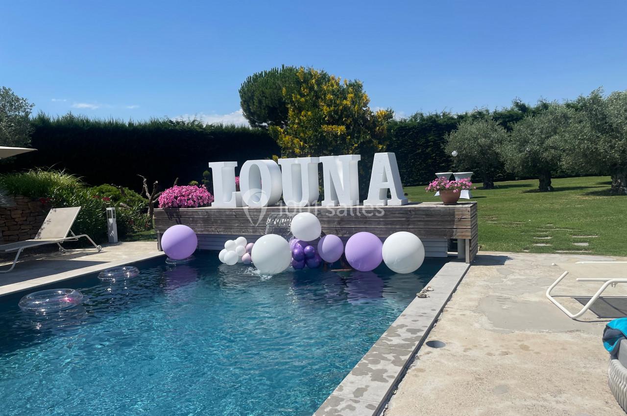 Piscine extérieure avec ballons blancs et violets flottants, décorée de grandes lettres formant le mot ’JOUNA’.