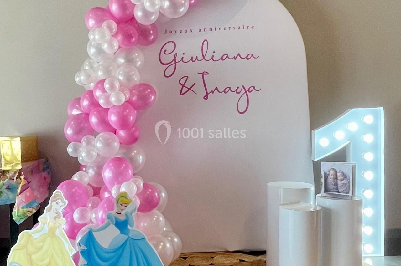 Décor d'anniversaire avec arche de ballons roses et blancs, silhouettes de princesses et chiffre lumineux ’1’.