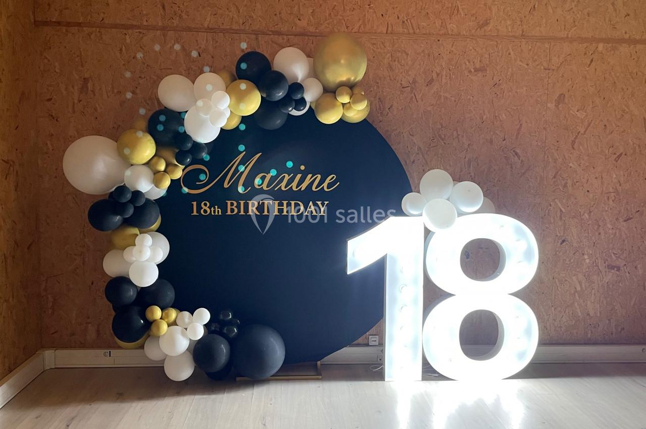 Décoration d'anniversaire avec un panneau noir orné de ballons et un chiffre lumineux ’18’ en blanc.