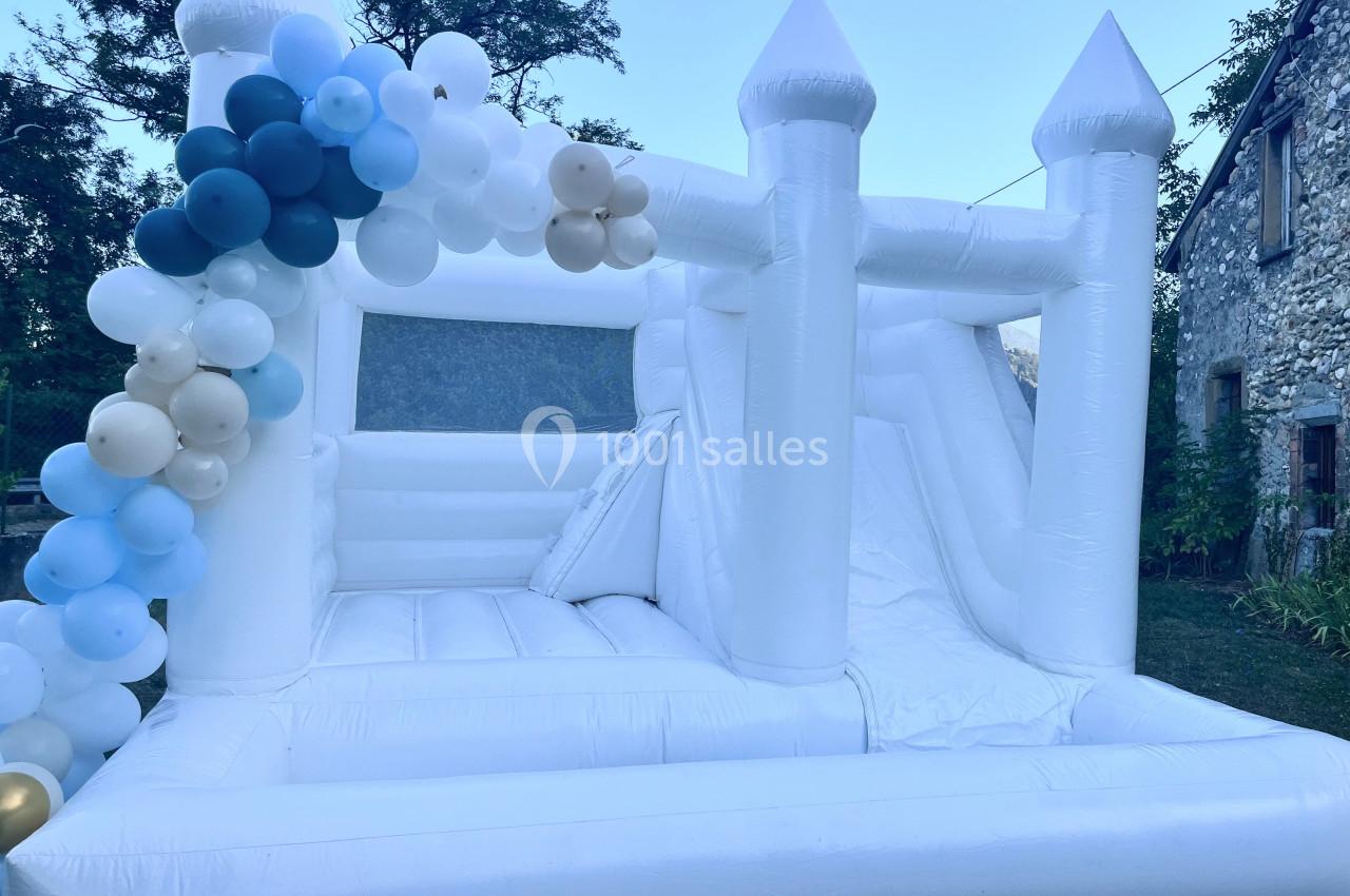 Château gonflable blanc avec toboggan, décoré de ballons colorés, installé dans un jardin près d'une maison en pierre.