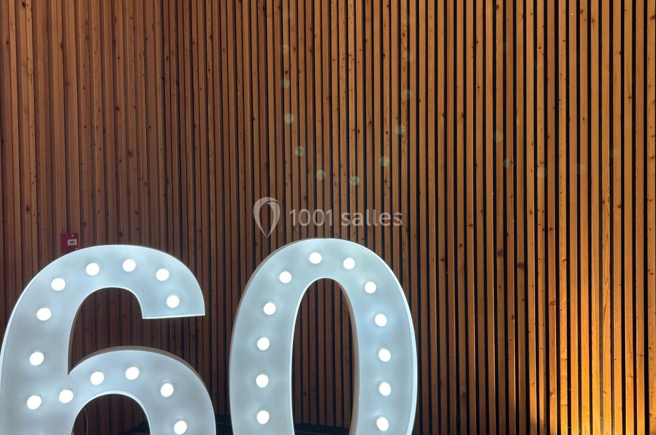 Chiffres lumineux ’60’ posés sur un sol en bois, devant un mur en lambris vertical.