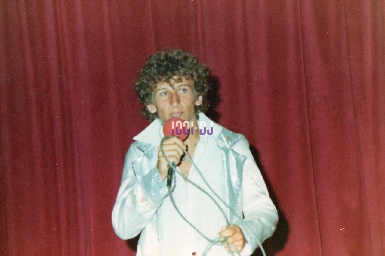 Un homme en chemise brillante chante sur scène devant un rideau rouge, tenant un micro à la main.