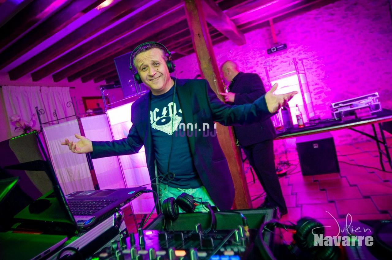 Un DJ souriant anime une soirée dans une salle éclairée par des lumières colorées, avec une table de mixage au premier plan.