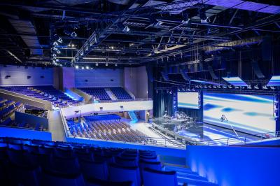 Vue aérienne d'un grand bâtiment moderne avec un parking rempli de voitures à l'avant et des arbres en arrière-plan.