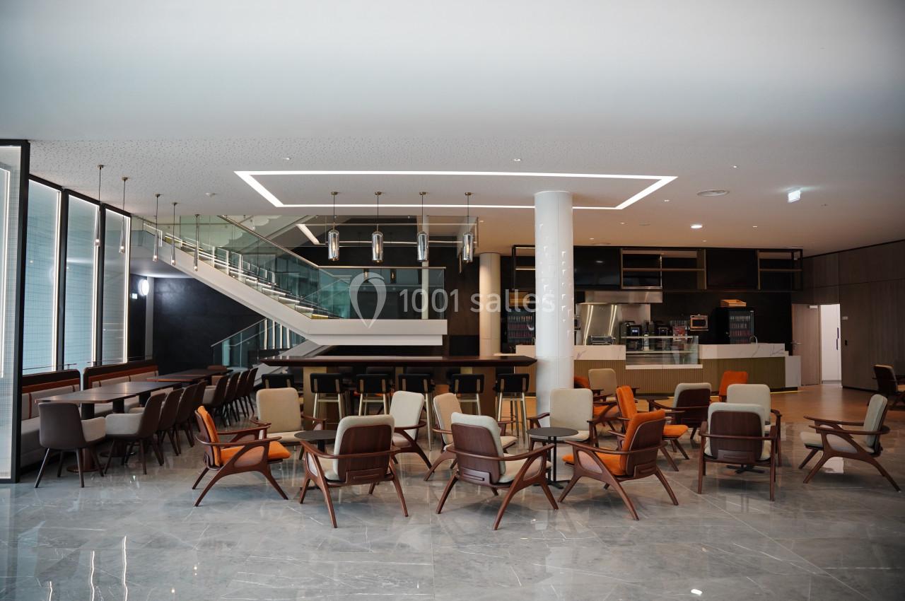 Salle moderne avec des chaises et tables disposées autour d'un espace ouvert, escalier et cuisine en arrière-plan.