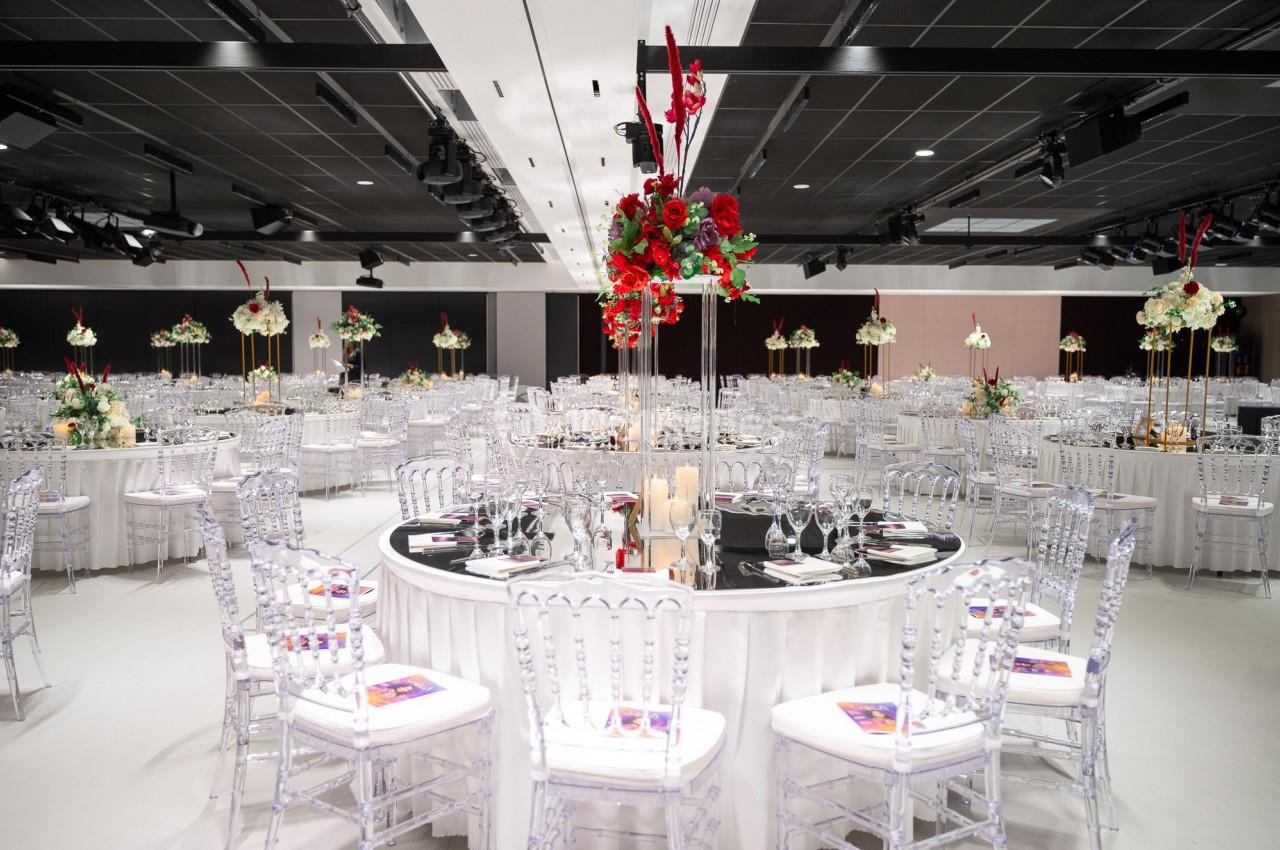 Salle de réception élégante avec tables rondes décorées de centres de table floraux rouges et blancs, chaises transparentes.