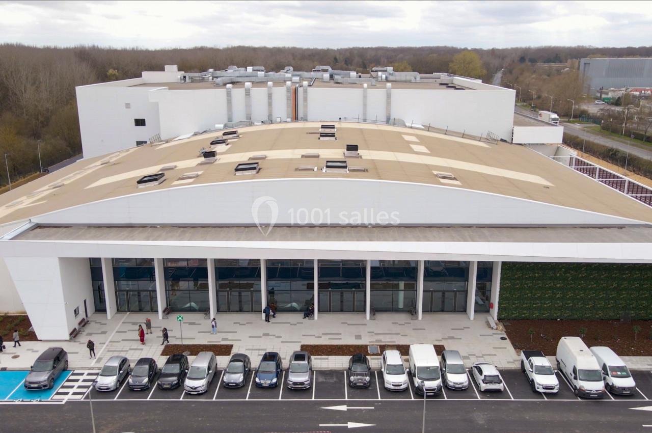 Vue aérienne d'un grand bâtiment moderne avec un parking rempli de voitures à l'avant et des arbres en arrière-plan.