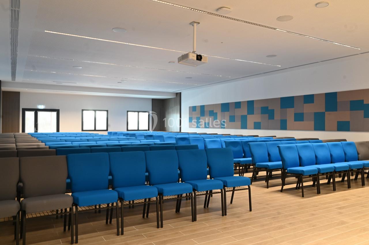 Salle de conférence moderne avec rangées de chaises bleues et grises, éclairée par la lumière naturelle et artificielle.