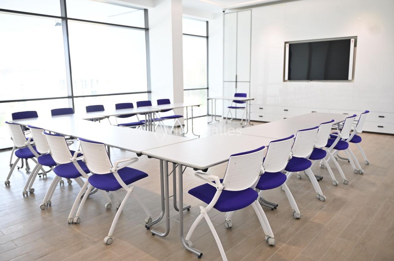 Salle de réunion lumineuse avec tables blanches disposées en U et chaises à assise violette sur un sol en bois clair.