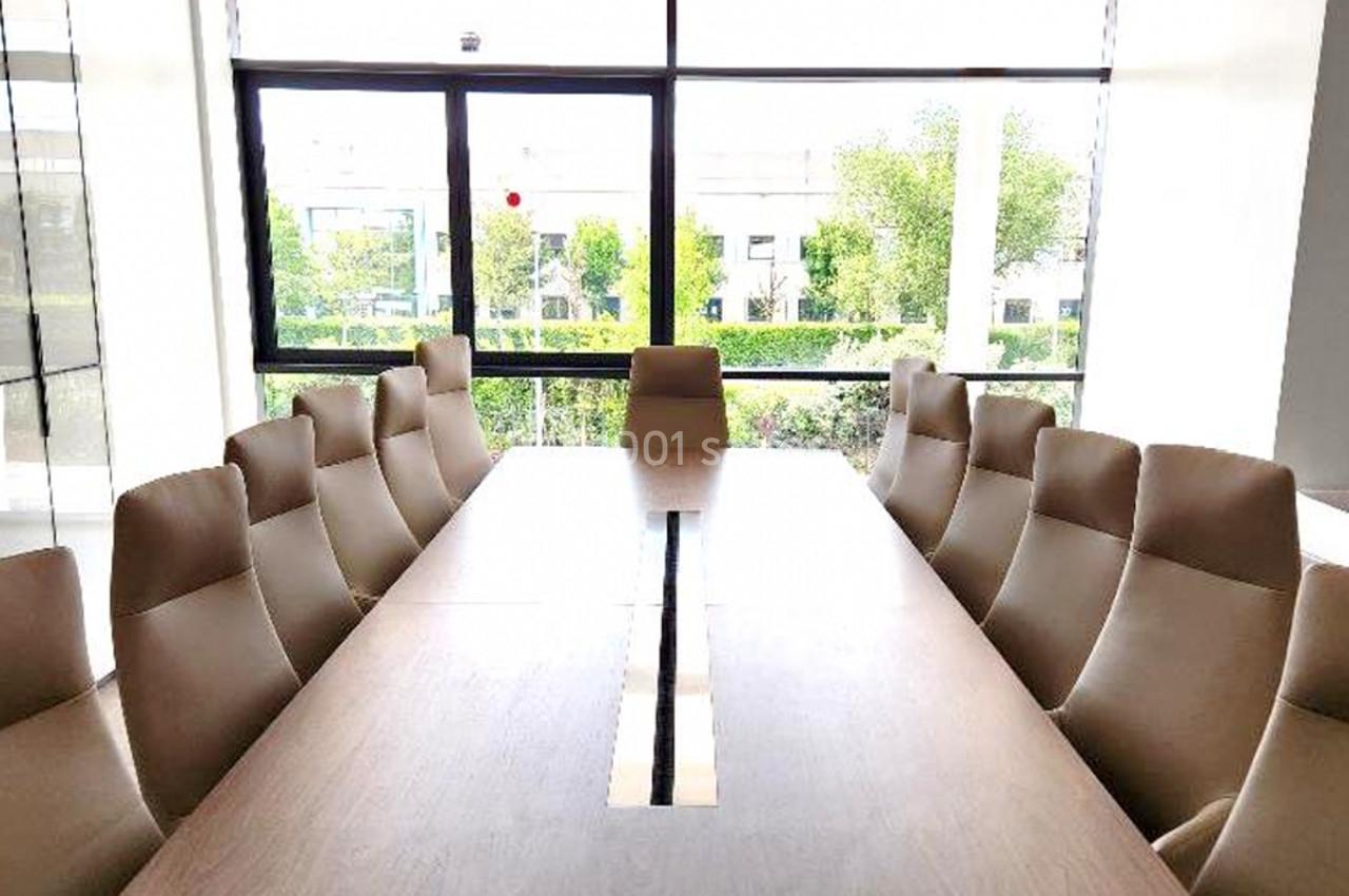 Salle de réunion lumineuse avec une grande table en bois entourée de chaises beige, vue sur des fenêtres donnant sur un…