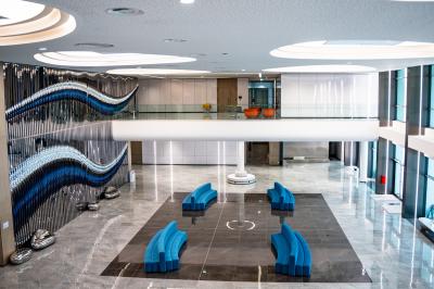 Vue aérienne d'un grand bâtiment moderne avec un parking rempli de voitures à l'avant et des arbres en arrière-plan.