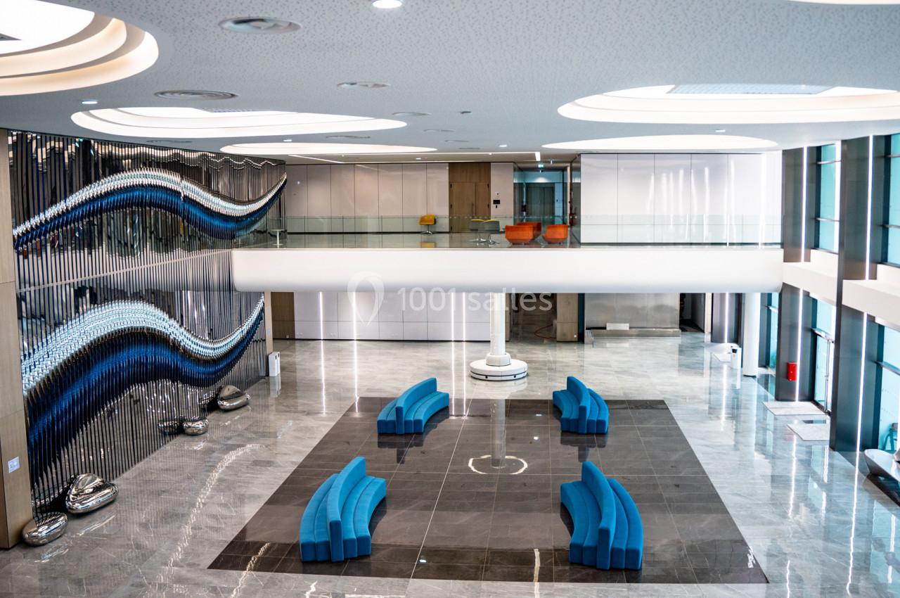 Hall moderne avec sol en marbre, sièges bleus incurvés, sculpture murale ondulée et mezzanine vitrée.