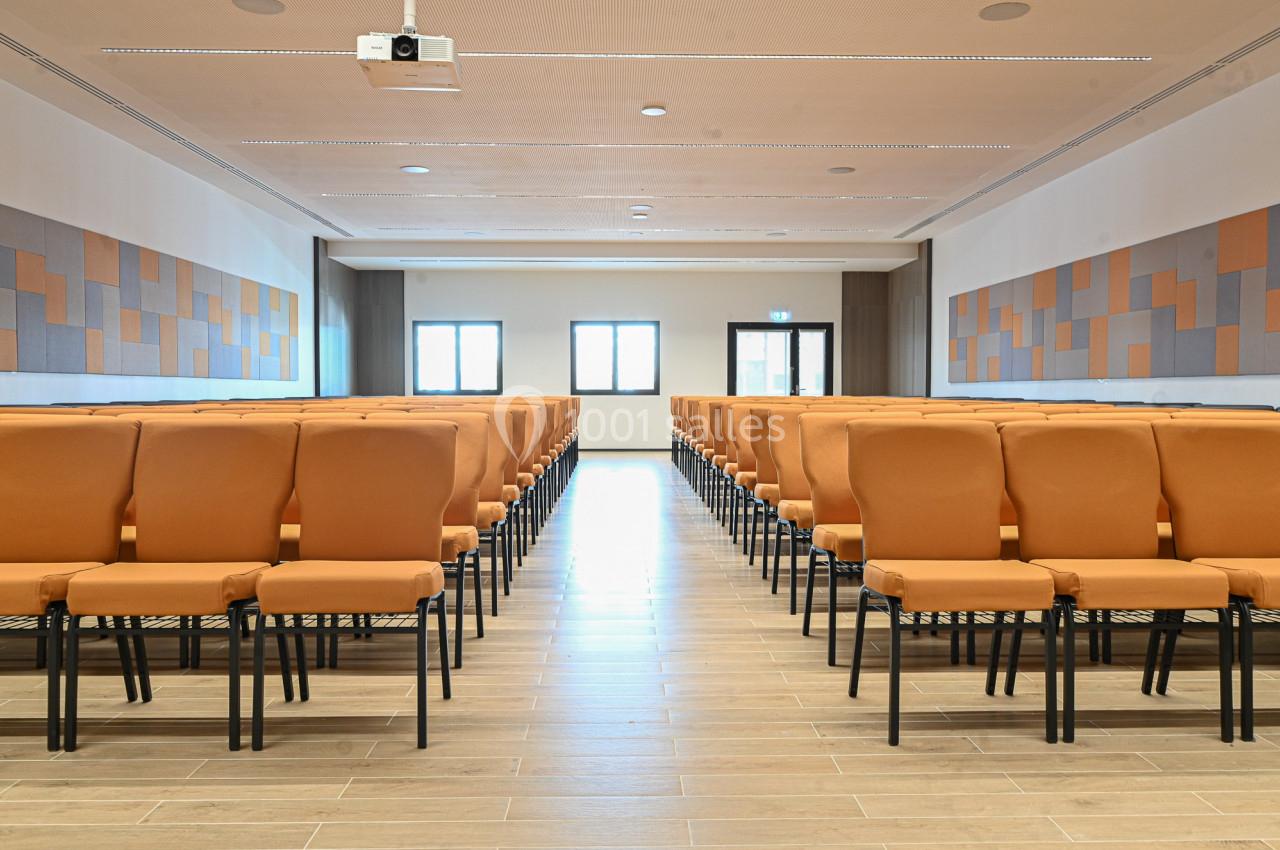 Salle de conférence lumineuse avec des rangées de chaises orange alignées face à un mur équipé d'un projecteur.
