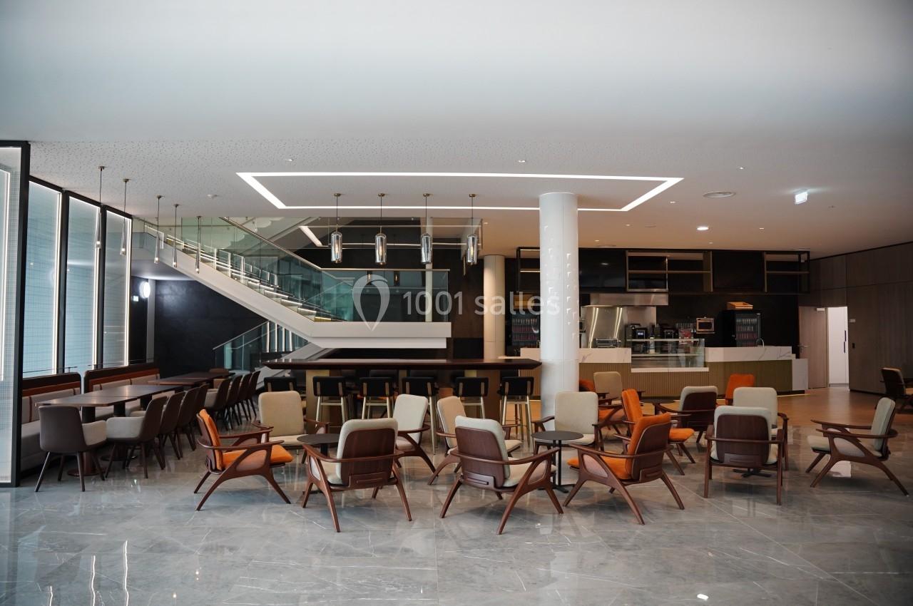 Salle moderne avec des tables, des chaises en bois et tissu, un escalier en arrière-plan et un espace cuisine ouvert.