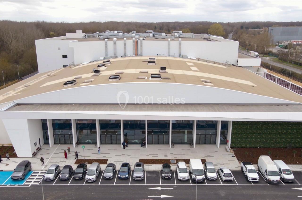 Vue aérienne d'un grand bâtiment moderne avec un toit beige, un parking rempli de voitures et des arbres en arrière-plan.