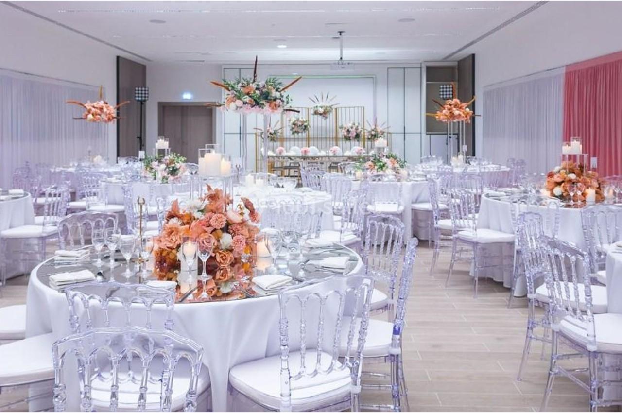 Salle de réception décorée avec des tables rondes, nappes blanches, chaises transparentes et arrangements floraux roses.