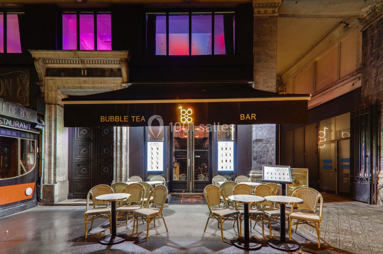 Façade d'un bar à bubble tea avec terrasse extérieure, tables et chaises en osier, éclairage nocturne.