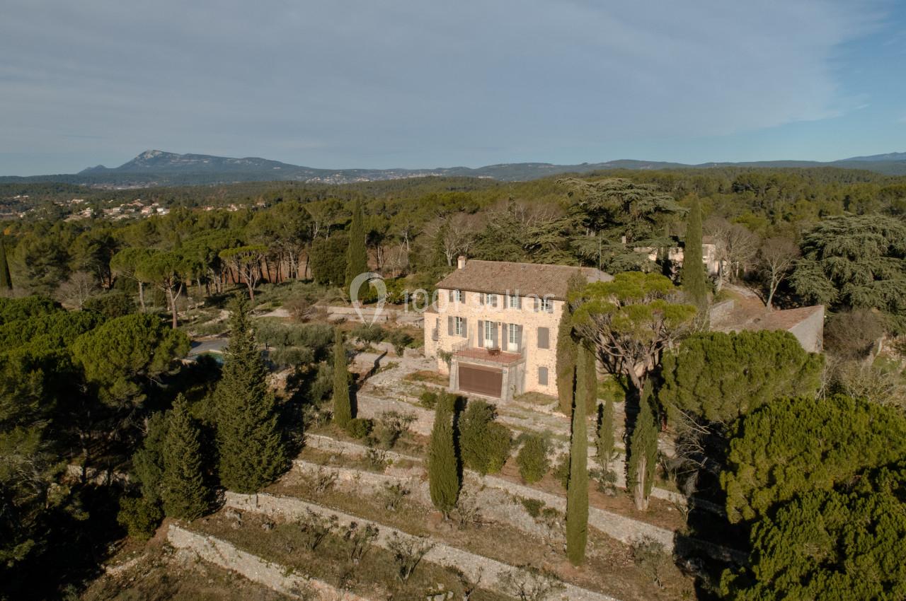 Vue aérienne d'une maison en pierre entourée de terrasses arborées et de végétation, avec des collines en arrière-plan.