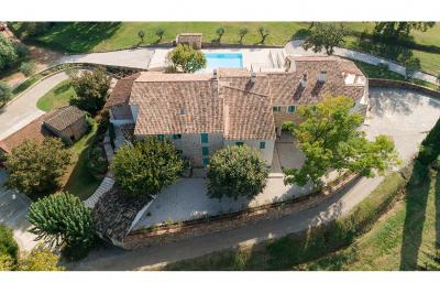 Vue aérienne d'une maison en pierre avec toit en tuiles, entourée d'arbres, d'une piscine et d'un chemin en gravier.