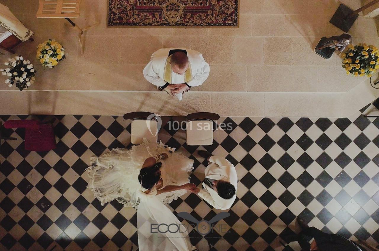 Vue aérienne d'un couple en tenue de mariage devant un officiant, sur un sol en damier noir et blanc.