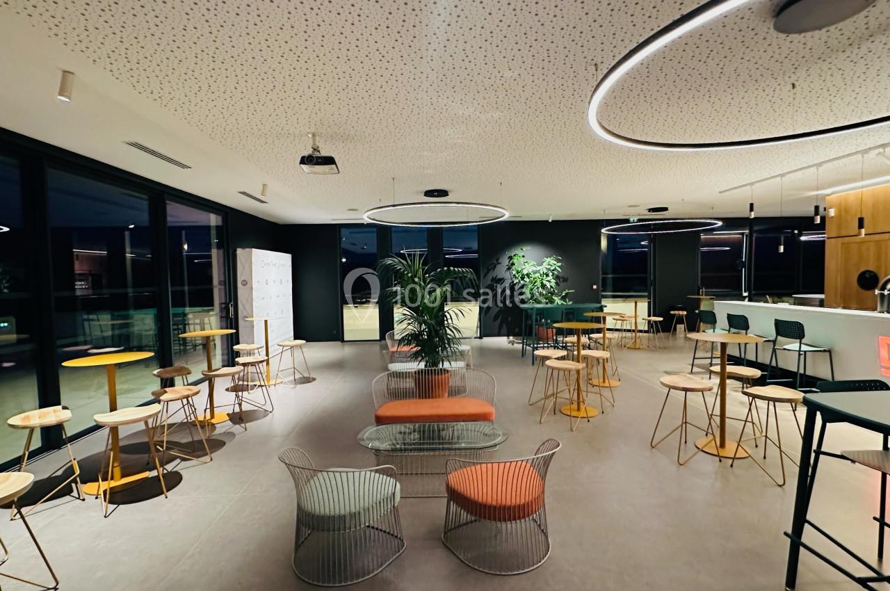 Salle moderne avec tables, chaises, plantes et éclairage circulaire, vue sur un comptoir et de grandes baies vitrées.