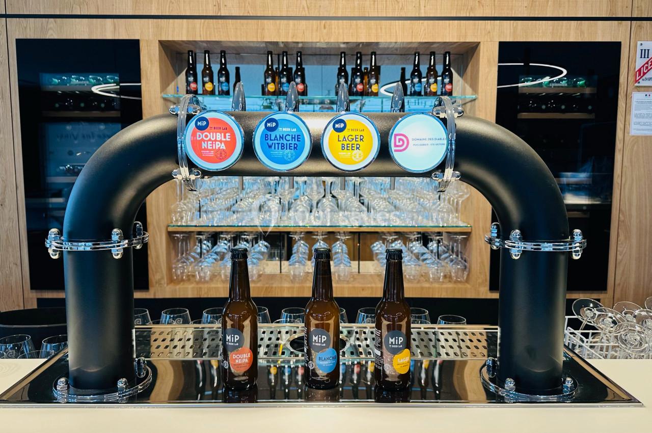 Tireuse à bière avec quatre robinets et bouteilles alignées devant, dans un bar moderne avec étagères de verres.