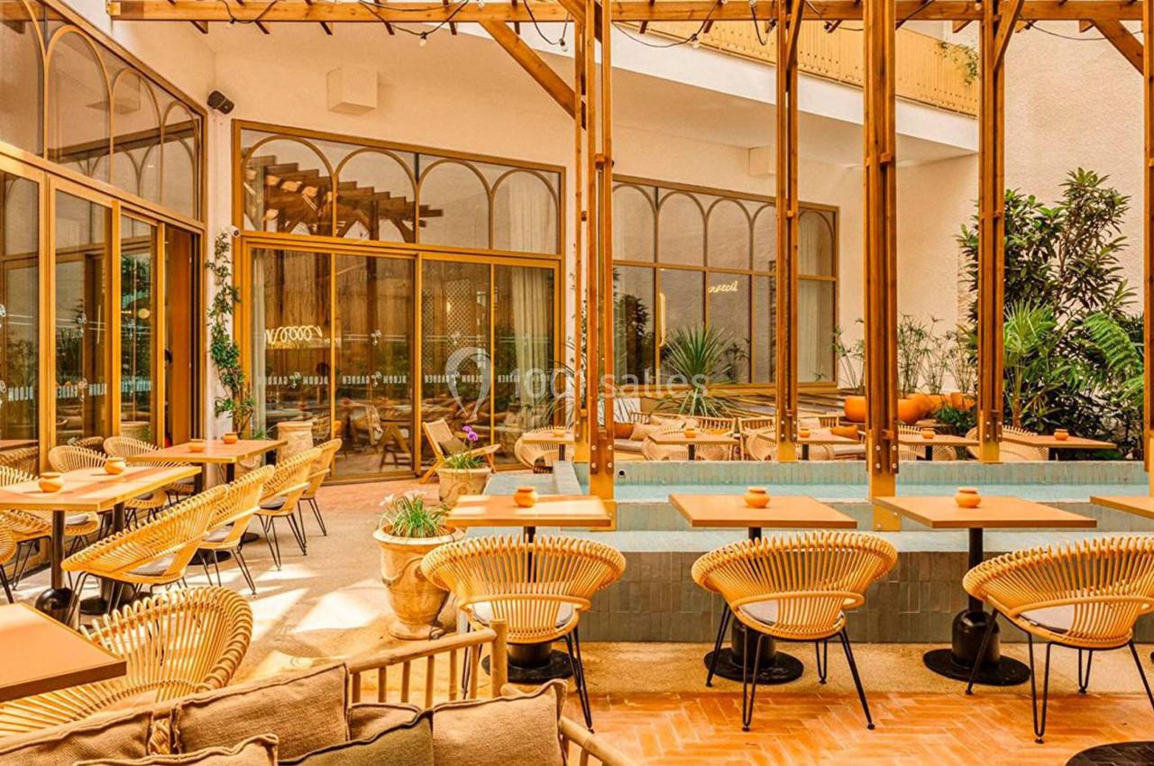 Terrasse lumineuse avec tables en bois, chaises en rotin et plantes, entourée de grandes baies vitrées.