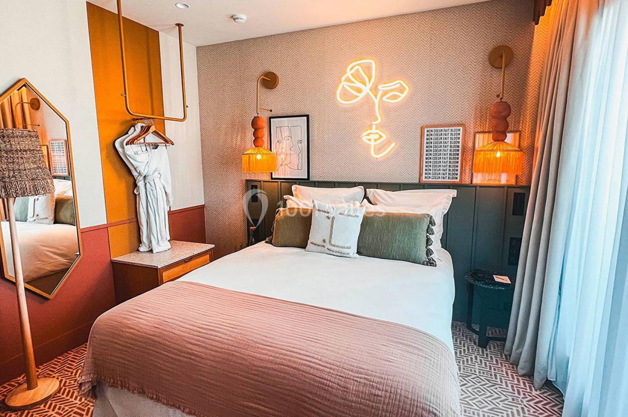 Chambre décorée avec lit double, statue, miroir, éclairage mural néon et rideaux laissant entrer la lumière naturelle.