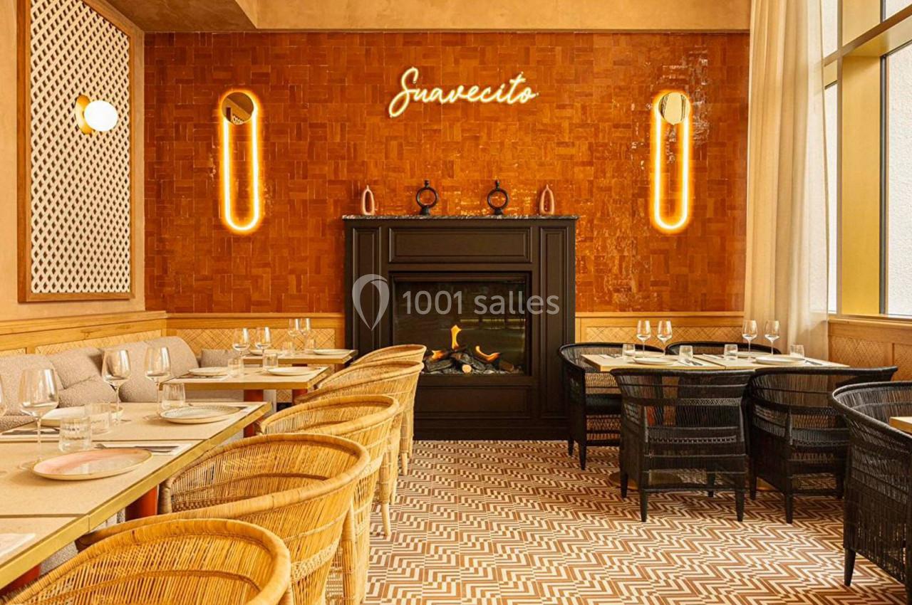 Salle de restaurant chaleureuse avec cheminée, murs en bois, néon ’Suavecito’ et mobilier en rotin et osier.