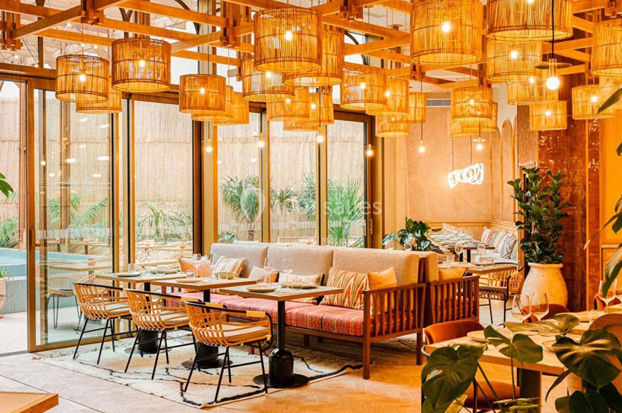 Salle de restaurant lumineuse avec mobilier en bois, luminaires suspendus et grandes baies vitrées donnant sur l'extérieur.